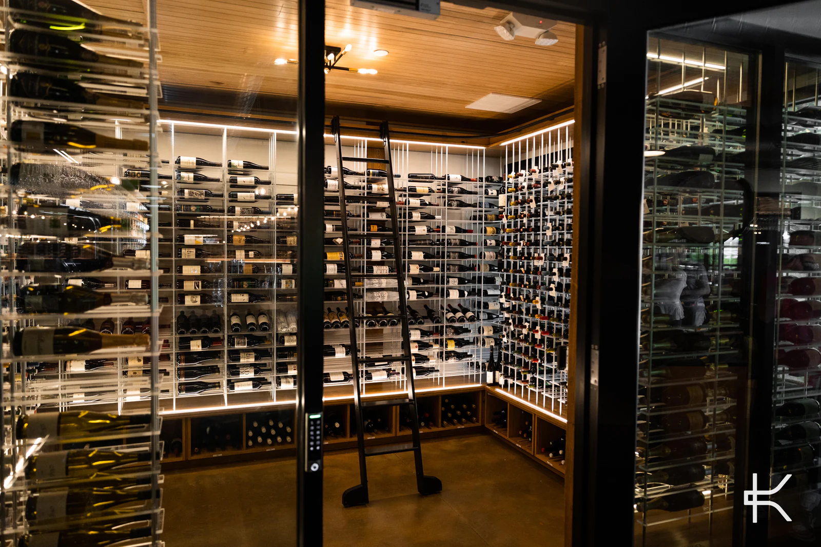 A glass wine cellar seen through black steel mullions, racks lit from below.
